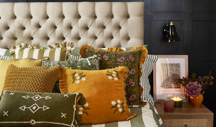 A cosy bedroom featuring a tufted beige headboard against dark panelling, styled with layered cushions in mustard yellow, olive green, and patterned fabrics. A striped duvet and floral throw add texture, while a bedside table with a black lamp, framed artwork, and lit candles create a warm, autumnal atmosphere.